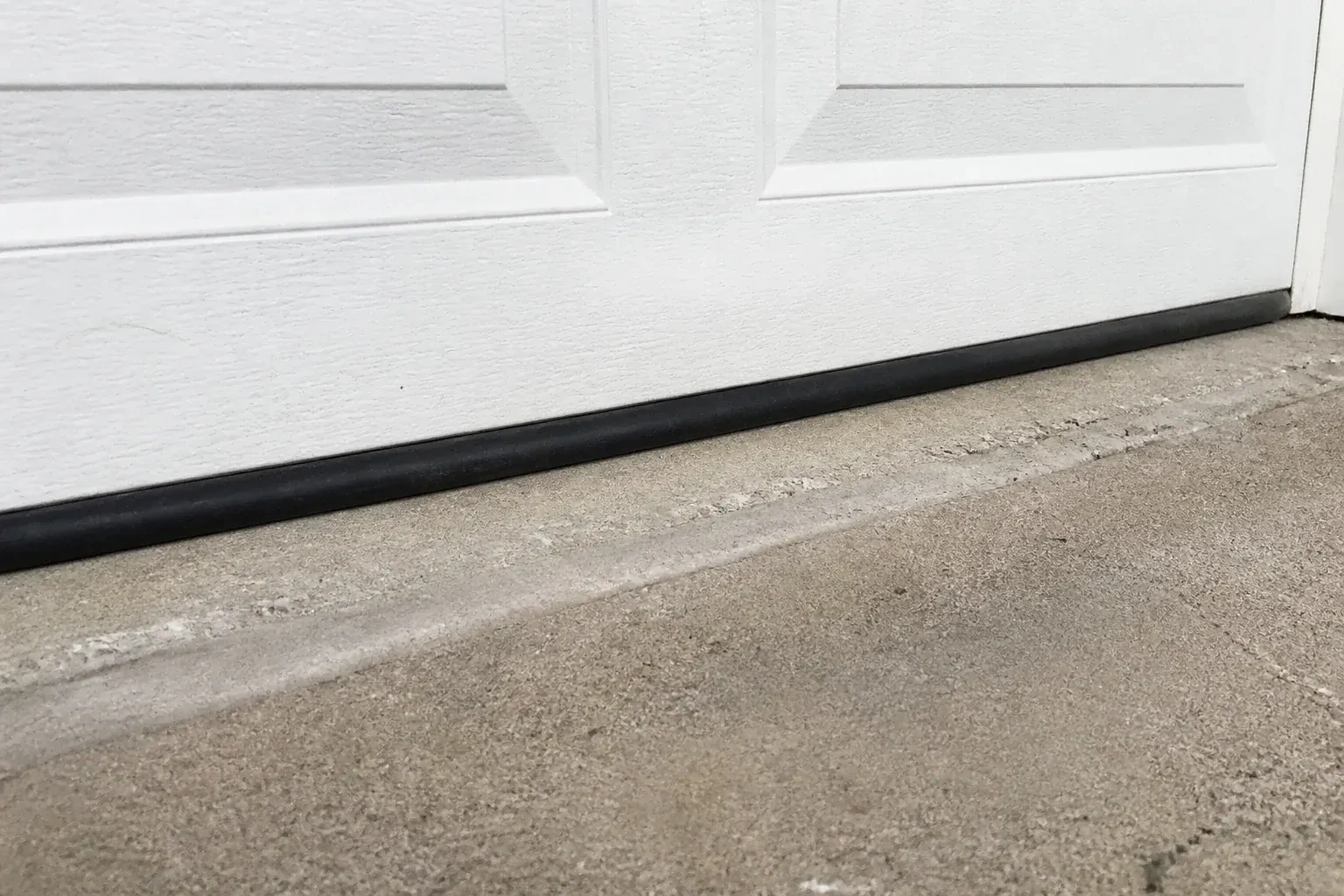 Black bottom seal along a closed white garage door at a concrete threshold. — Garage Door Bottom Seal Replacement, Kitchener